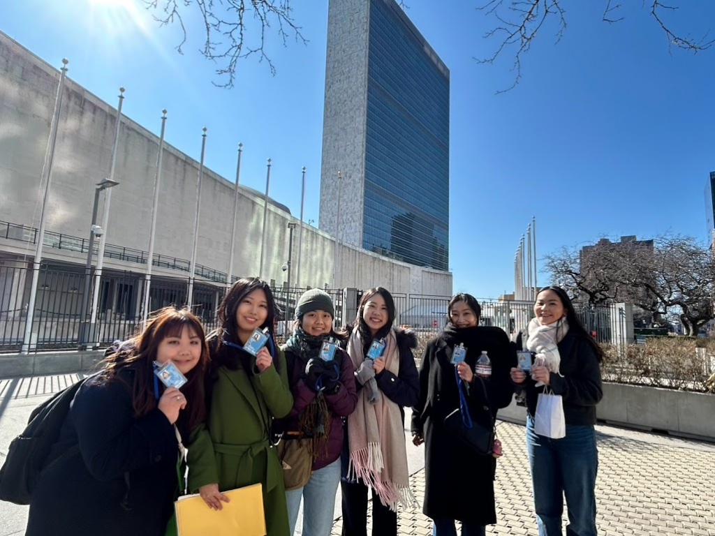 Six young women smile for a photo outdoors in front of the United Nations Headquarters building on a sunny day. They are dressed for cool weather and are holding up their event ID badges toward the camera.