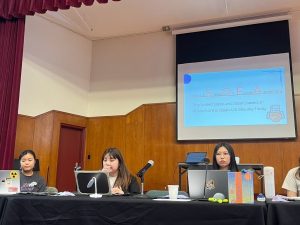 Three young women sit as panelists behind a table with laptops and a microphone. Behind them, a presentation slide is projected with the title "Japan-U.S. SOFA (Status of Forces Agreement)" and a description related to the Japan-U.S. Security Treaty.